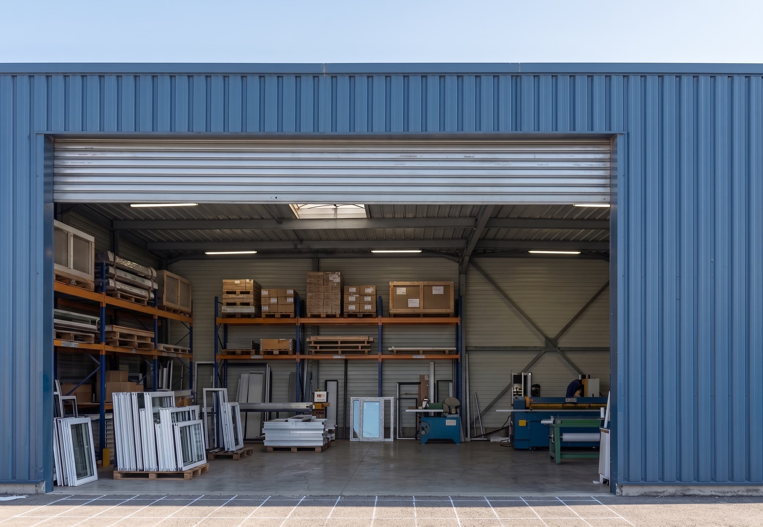 Intérieur de l'atelier Équipe Baie vu depuis la porte : stock de profilés, machines et commandes palettisées prêtes au départ
